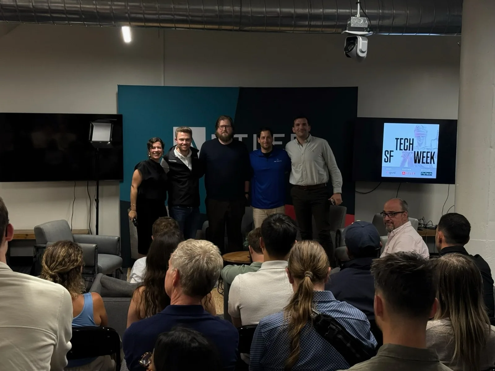 The moderator and four panelists stand together in front of the Antler banner, beside a screen showing the SF Tech Week program.